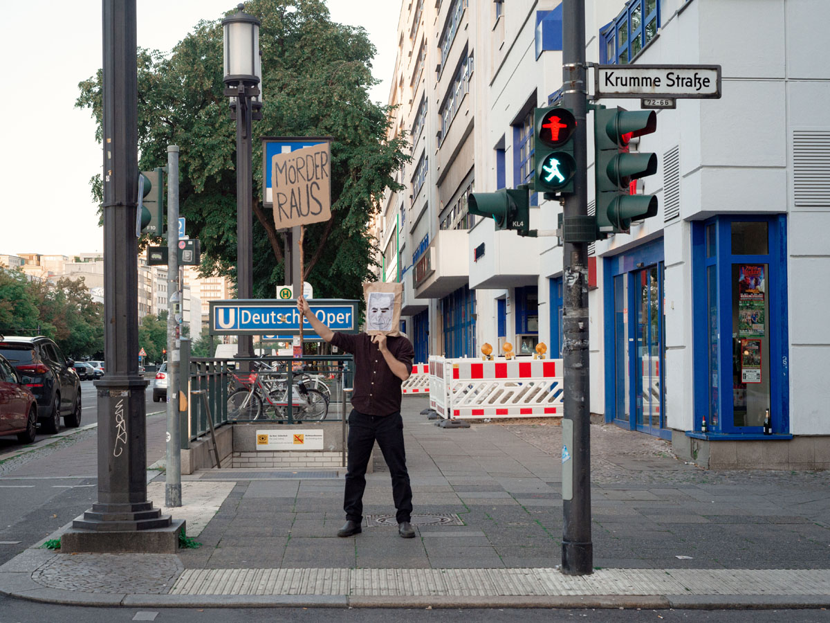 Foto mit einer Person auf der Straße, die eine Papiertüte auf dem Kopf hat und ein Banner mit der Aufschrift Mörder raus hochhält. Foto von Alex Bex